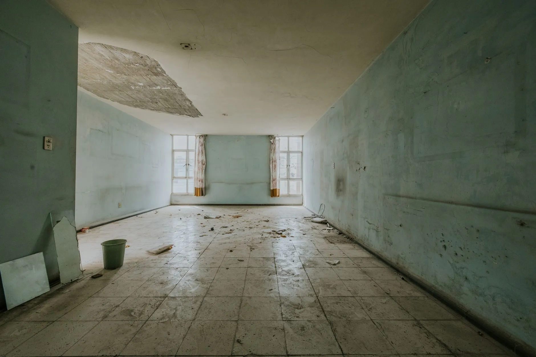 An empty room with a section of ceiling plaster fallen after a leak, a bucket on the floor