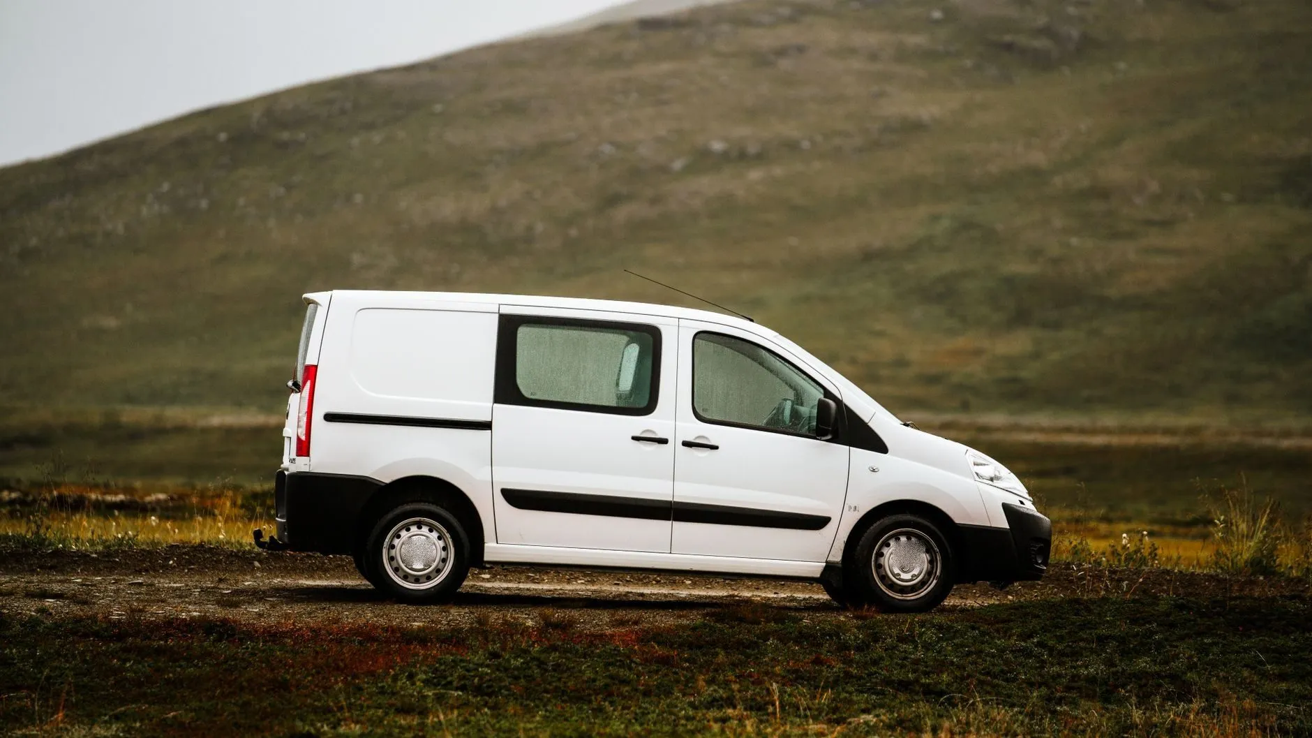 A plain white panel van parked on a gravel track below a green hillside