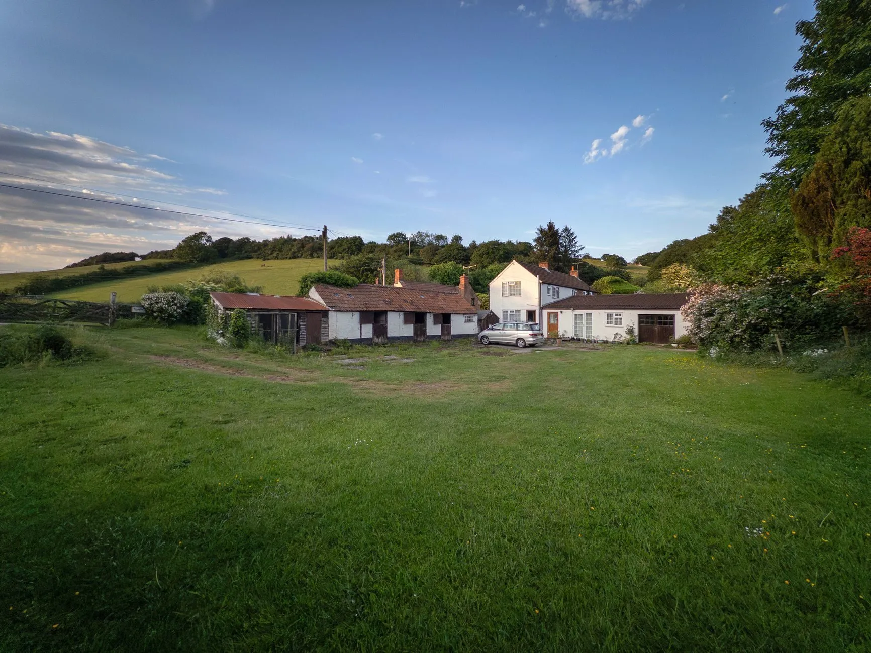 A white farmhouse, outbuildings and a yard below a wooded hillside