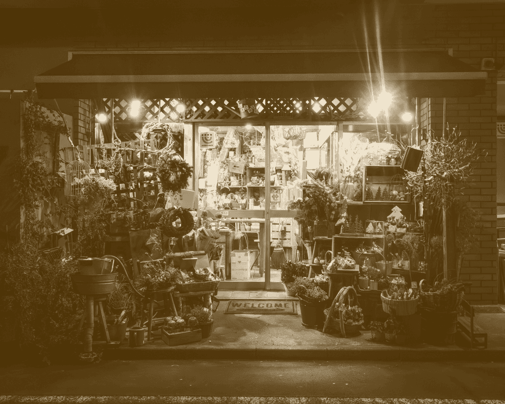 An independent shopfront lit at night, its stock out on the pavement, printed in one ink.